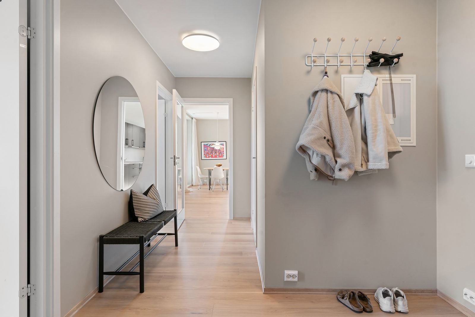 Bright entryway hallway with a wall-mounted hook rail holding coats, a round mirror, a bench and shoes on the floor